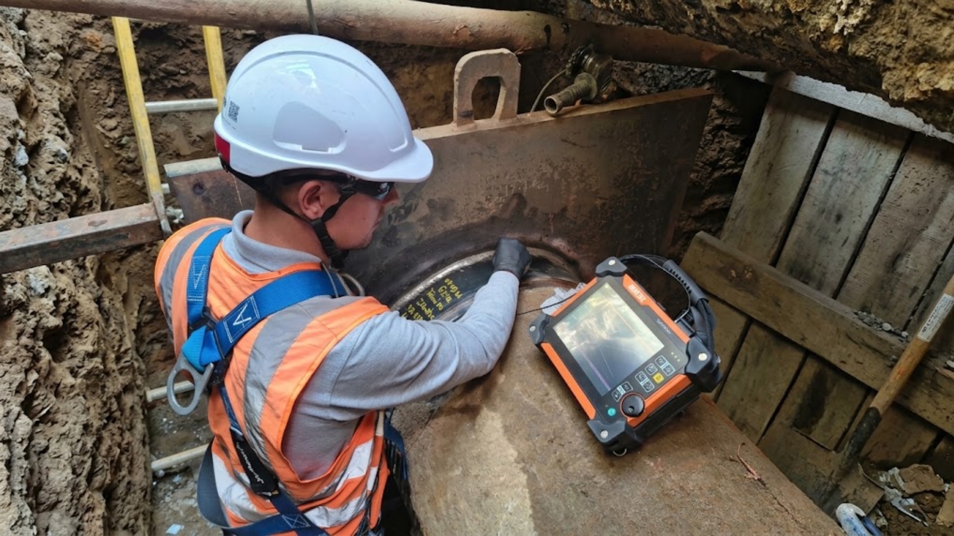 Técnico con arnés inspeccionando por ultrasonido una tubería dentro de una excavación